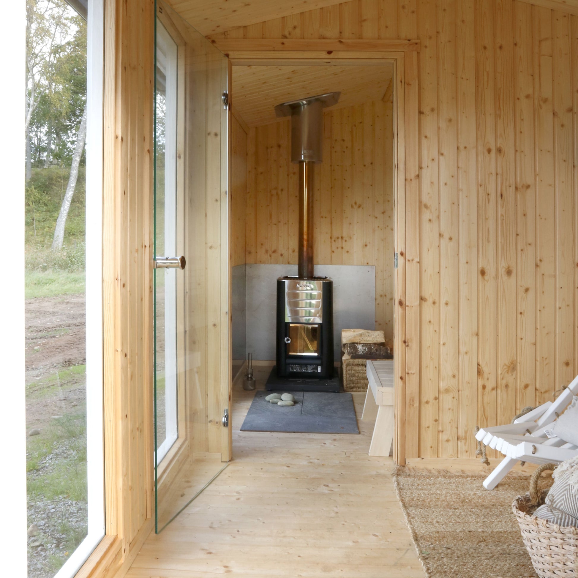 Interiör av en bastustuga i trä med glasdörr, vedeldad kamin, sittbänkar och vedförvaring. Ljus och naturlig atmosfär med utsikt mot grönska utanför.